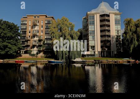 Moderni blocchi di appartamenti e un grande salice piangente riflesso in un canale tranquillo con piccole barche, sotto il cielo blu in una zona residenziale di Amsterdam. Foto Stock
