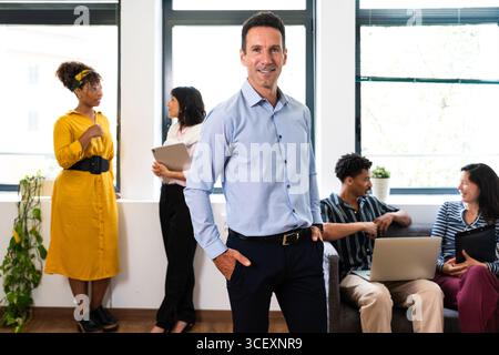 Un uomo d'affari sicuro di sé sta con le mani in tasca, conducendo una riunione di team diversificata in un ufficio luminoso e moderno, favorendo la collaborazione e l'innovazione Foto Stock