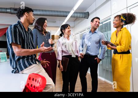 Cinque persone d'affari allegri e diversificati stanno organizzando riunioni produttive, scambiando idee e sorridendo mentre si trovano in un moderno corridoio di uffici, ho Foto Stock