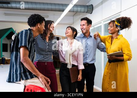 Cinque allegri colleghi d'affari multietnici che si abbracciano e ridono insieme in un ufficio luminoso e moderno, celebrando il successo del loro progetto e del loro team Foto Stock