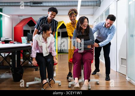 I membri del team aziendale di startup si spingono a vicenda su sedie rotanti e ridono in un ambiente di ufficio moderno, godendosi una pausa dal lavoro e dalle esibizioni Foto Stock
