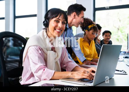 Team aziendale di startup che collabora e lavora al progetto in un ufficio moderno, un rappresentante del servizio clienti che indossa le cuffie e sorride durante la digitazione Foto Stock