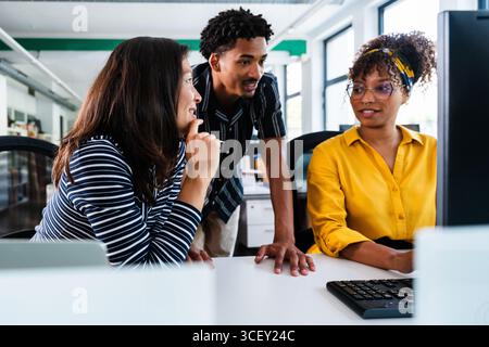 Tre diversi colleghi lavorano insieme in un ufficio moderno, impegnandosi in un lavoro di squadra produttivo mentre analizzano i dati sullo schermo di un computer Foto Stock