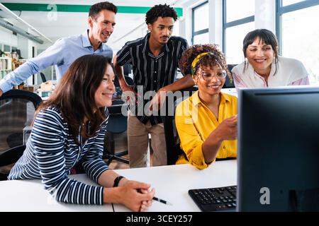 Diversi gruppi di professionisti si sono riuniti intorno a un computer, discutendo un progetto e condividendo idee in un ambiente d'ufficio luminoso e moderno, dimostratina Foto Stock