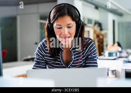 Donna d'affari concentrata che indossa una cuffia/auricolare partecipa a una videochiamata mentre lavora in remoto dal suo ufficio domestico, utilizzando un laptop per una c Foto Stock