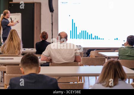 Seminario con partecipanti che ascoltano un oratore che presenta un grafico su uno schermo in una moderna sala conferenze. Evidenziando il lavoro di squadra, la condivisione dei dati e le dinamiche di sviluppo professionale. Foto Stock