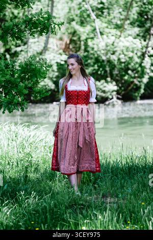 Un ritratto a figura intera di una bella giovane donna con lunghi capelli marroni, che indossa un tradizionale dirndl rosso con una camicetta bianca e un assegno rosso e bianco Foto Stock