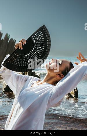 Giovane ballerina di flamenco in posa drammatica sul mare con ventilatore nero Foto Stock