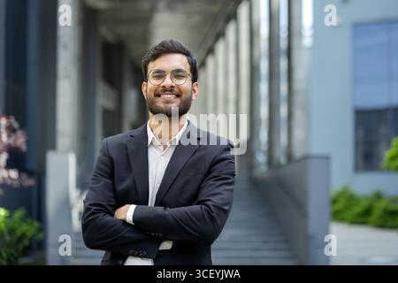 Un uomo d'affari sorridente che indossa un abito e un paio di occhiali si posa in modo sicuro in un ambiente urbano all'aperto. Foto Stock