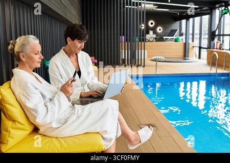 Due donne adorabili si rilassano in piscina, godendosi del tempo libero con i gadget in un centro benessere. Foto Stock
