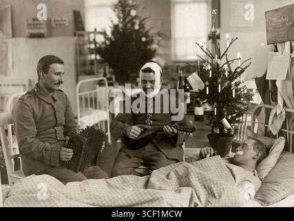 Prima guerra mondiale, 1914: Un soldato ferito giace in un letto d'ospedale a Berlino. Un altro soldato, ferito alla testa, si siede accanto a lui, mentre un uomo suona l'armonica. Un piccolo albero di Natale decorato si erge accanto al letto, con uno più grande più in basso nella camera. Foto Stock