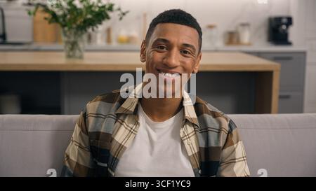 Ritratto a casa sorridente ragazzo maschio schietto giovane calmo che riposa un uomo etnico afroamericano in salotto che guarda la macchina fotografica sul divano da solo Foto Stock