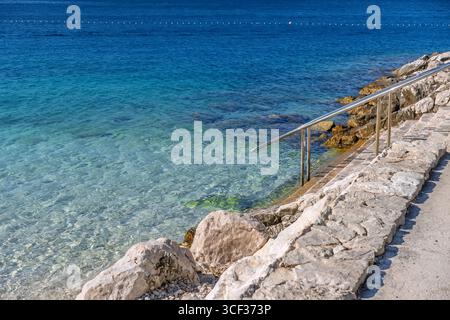 Invitante vista di una tranquilla baia mediterranea con una costa rocciosa e un corrimano per un facile accesso al mare. Spiaggia panoramica con acque cristalline e azzurre Foto Stock