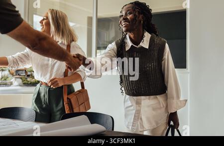 Due architetti donna, una giovane adulta e una di mezza età, stringono la mano a un cliente nel loro ufficio moderno, simboleggiando il lavoro di squadra e la collaborazione professionale Foto Stock