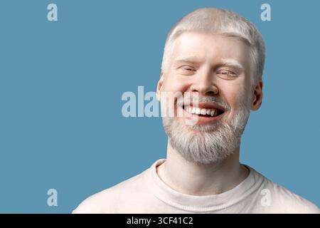 Ritratto di un uomo albino barbuto felice con pelle pallida e capelli bianchi che sorride alla fotocamera su sfondo turchese Foto Stock