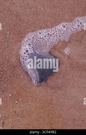 Una pietra a forma di cuore in sabbia bagnata, schiuma, spray Foto Stock