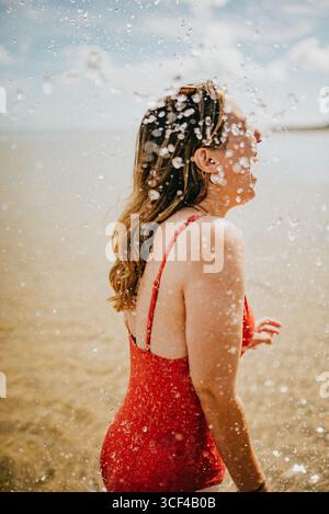Una donna con un costume da bagno rosso è in piedi in acqua, spruzzando acqua su se stessa. Concetto di divertimento e giocosità, mentre la donna si sta divertendo Foto Stock