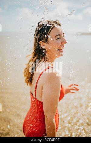 Una donna con un costume da bagno rosso spruzza acqua su se stessa mentre sorride. Concetto di divertimento e giocosità, mentre la donna si sta divertendo in acqua Foto Stock