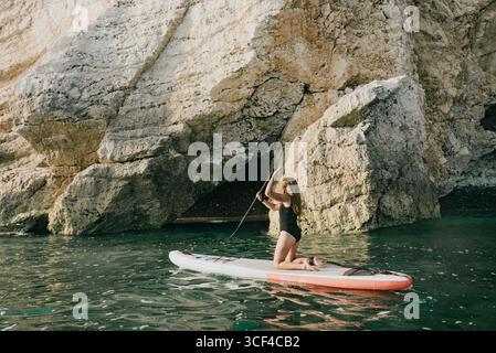 Una donna sta pagaiando su una tavola da surf in un corpo d'acqua. L'acqua è calma e la donna è in bikini nero Foto Stock