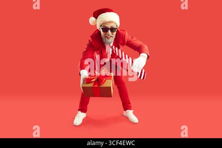 Uomo in tuta rossa e cappello di Babbo Natale in piedi su sfondo rosso e ti regala un regalo di Natale Foto Stock