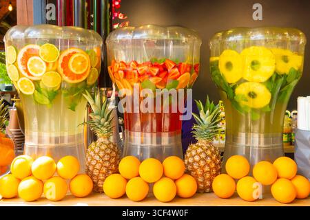 Tre dispenser trasparenti con bevande a base di frutta fresca, tra cui limone, fragole e ananas, con arance intere e ananas su un tavolo di legno Foto Stock