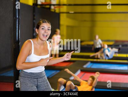 Felice giovane donna europea in cima bianca e leggings grigi che si posano sui trampolini durante il fine settimana attivo tempo libero nel centro di intrattenimento Foto Stock