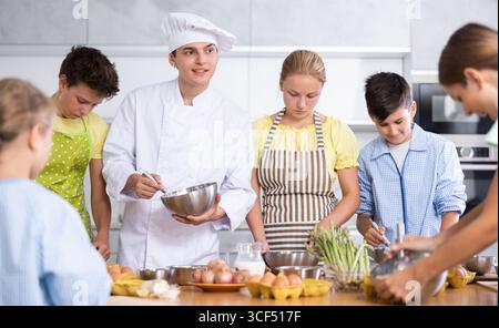 Durante la lezione di cucina, il ragazzo cuoco con la ciotola in mano racconta ai bambini partecipanti zecche e trucchi culinari Foto Stock
