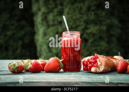Marmellata di fragole fatta in casa in un barattolo con baguette fresca su un vecchio tavolo di legno all'aperto. Spalmabile a base di frutta fresca di stagione. Primo piano. Foto Stock