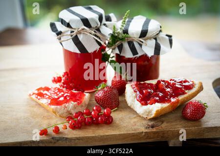 Natura morta con marmellata fatta in casa di fragole e ribes in vaso. Frutta rossa e baguette con marmellata appena cotta su tagliere rustico in legno. Foto Stock