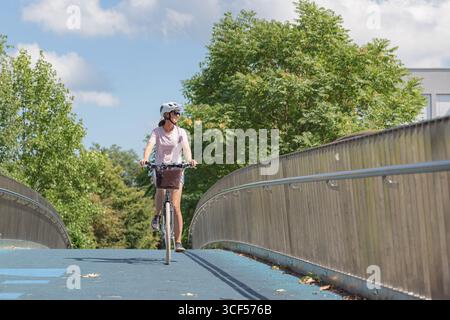 Una giovane donna cavalca la sua bicicletta su un ponte moderno circondato da alberi lussureggianti sotto un cielo azzurro cristallino durante il pomeriggio. Foto Stock