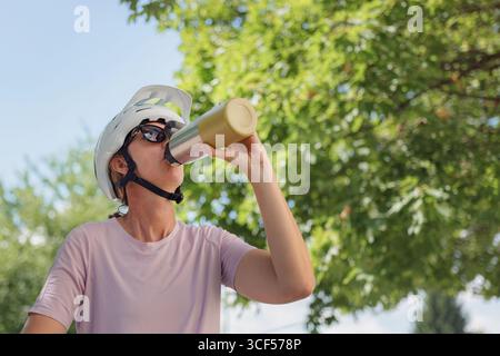 Una donna che indossa un casco beve da una bottiglia di acciaio inossidabile mentre si gode l'ombra degli alberi durante la sua pausa ciclistica. Foto Stock