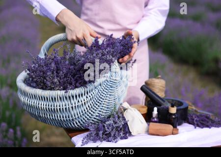 Fare cosmetici naturali. Donna con cesto in vimini di fiori di lavanda e altri ingredienti a tavola nel campo, primo piano Foto Stock