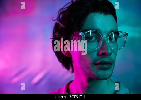 Foto in primo piano orizzontale di un bellissimo giovane uomo caucasico che indossa occhiali da vista in piedi con luce al neon blu e rosa che guarda la fotocamera Foto Stock