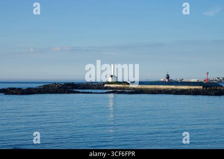 Un faro bianco sorge sulla frangiflutti del porto di Gwideok, fiancheggiato da fari luminosi lungo le mura del porto, mentre l'acqua blu calmo riflette la mattina Foto Stock