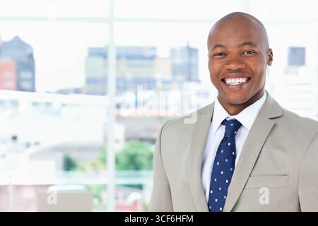 Uomo afroamericano in piedi davanti alla finestra in ufficio con vestito grigio con cravatta blu navy, copia spazio Foto Stock