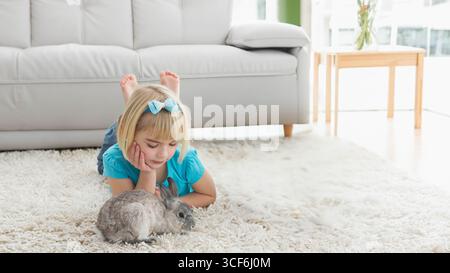 Bambina sdraiata su un tappeto d'avorio nel soggiorno che accarezza conigli grigi sotto la finestra, spazio copia Foto Stock