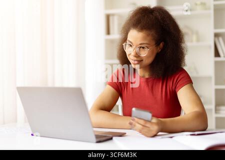 Giovane donna afroamericana con occhiali che lavora al computer portatile e che tiene in mano lo smartphone Foto Stock