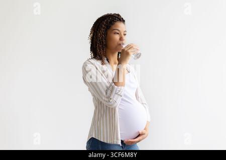 Donna afroamericana incinta che beve un bicchiere d'acqua e tocca la pancia Foto Stock
