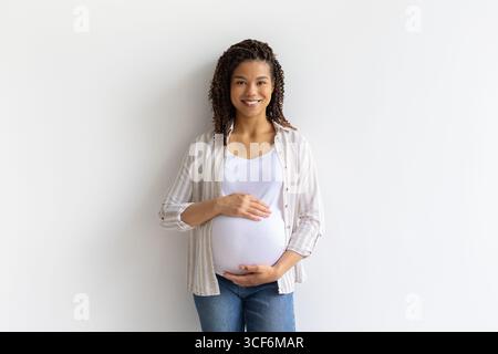 Sorridente donna nera incinta in piedi su sfondo bianco, tenendo la pancia con le mani Foto Stock