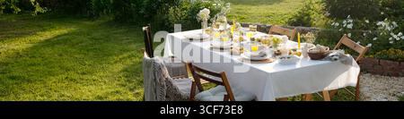 Tavolo da pranzo all'aperto con piatti, bicchieri, pane, insalate, succo di frutta fiori disposti su tovaglia, sedie di legno che circondano il tavolo in giardino con erba verde Foto Stock