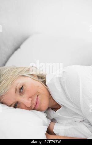 Donna che riposa la testa su un cuscino bianco e sorride alla macchina fotografica sul letto con piumone, spazio fotocopie Foto Stock