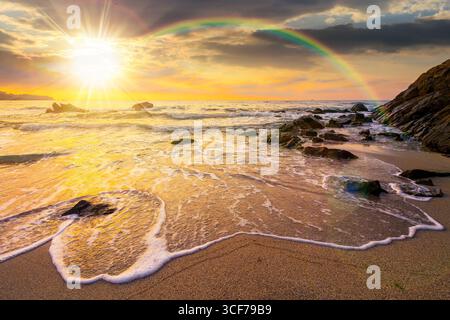 costa marina con arcobaleno sopra i massi nella baia al tramonto. la marea del mare con onde calme bagna la spiaggia di sabbia dorata con la luce della sera. bell'autunno Foto Stock
