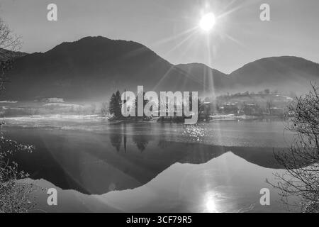 Vista invernale in bianco e nero del Lago d'Idro in Lombardia, con sole luminoso, montagne alpine e alberi che si riflettono su un'atmosfera d'acqua calma Foto Stock