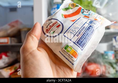 Baviera, Germania - 21 agosto 2025: Una mano tiene una mozzarella davanti a un frigorifero aperto. L'etichetta Nutri-Score mostra B. immagine simbolica per lo shopping, l'etichettatura degli alimenti e la nutrizione consapevole *** Eine Hand hält eine Mozzarella-Packung vor einem offenen Kühlschrank. DAS Nutri-Score-Label zeigt B. Symbolbild für Einkauf, Lebensmittelkennzeichnung und bewusste Ernährung Foto Stock