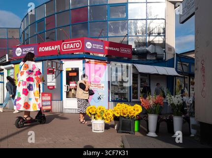 Kiev, Ucraina - 20 luglio 2025: Donna in sella a uno scooter elettrico davanti a un cavalletto floreale mentre un'altra donna parla al telefono di fronte a un pedone in Ucraina Foto Stock