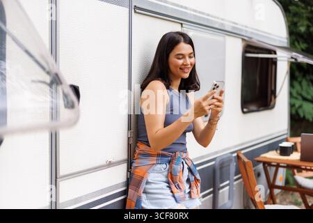 Donna felice in piedi vicino a un camper, che utilizza lo smartphone durante un viaggio in campeggio in campagna, copia spazio. Bella ragazza che naviga sul Web, chatta online, guarda Foto Stock