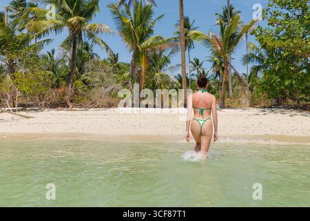 Attraente giovane donna in bikini che esce dall'acqua di mare su una splendida spiaggia sabbiosa con palme, godendo una vacanza sulla costa tropicale, sul retro Foto Stock