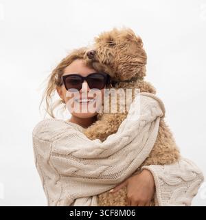 Una donna felice e sorridente che indossa gli occhiali da sole abbraccia il suo carino cane Cockapoo in una giornata ventosa sulla spiaggia di Saltburn-by-the-Sea, North Yorkshire, Regno Unito. Foto Stock