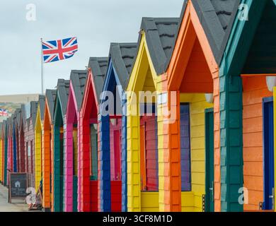 Le tradizionali capanne da spiaggia dipinte con colori vivaci fiancheggiano il lungomare, con una bandiera Union Jack che sventola, presso la località balneare di Saltburn, North Yorkshire, Regno Unito. Foto Stock
