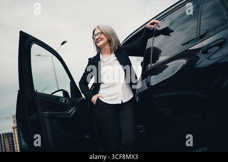 Elegante donna matura professionale che abbandona un'auto elegante in un ambiente urbano, mostrando eleganza e successo Foto Stock